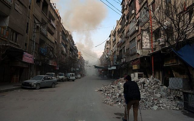 Un sirio con muletas camina por una calle mientras el humo se expande en la ciudad rebelde de Douma, en la sitiada región oriental de Ghouta, en las afueras de la capital, Damasco, tras los ataques aéreos de las fuerzas del régimen en la zona, el 23 de febrero de 2018. (AFP / HAMZA AL-AJWEH)