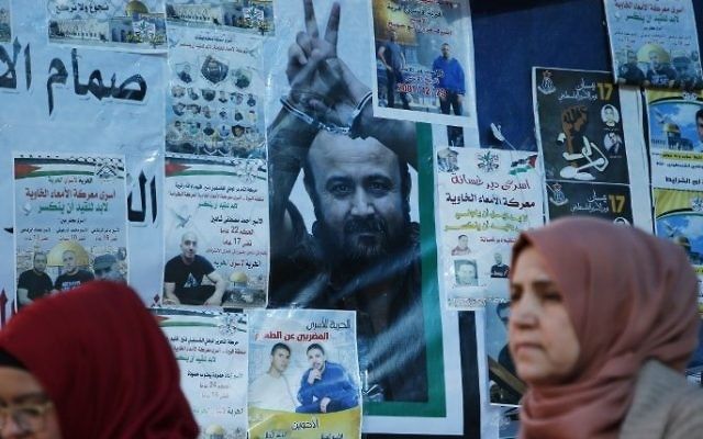 Mujeres palestinas pasan frente a un muro con afiches, incluido un retrato del architerrorista preso Marwan Barghouti, durante una manifestación en Ramallah, en apoyo de él y otros terroristas presos en huelga de hambre en las cárceles israelíes el 24 de abril de 2017. (AFP / Abbas Momani)