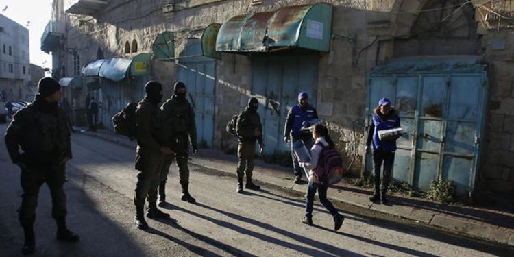 Observadores palestinos, a la derecha, observan cómo los niños pasan junto a los soldados israelíes en su camino a la escuela en la ciudad cisjordana de Hebrón