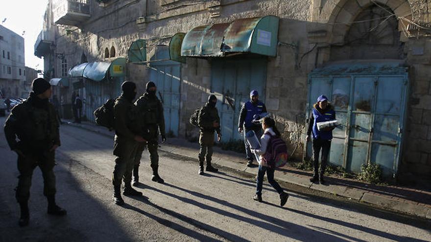 Observadores palestinos, a la derecha, observan cómo los niños pasan junto a los soldados israelíes en su camino a la escuela en la ciudad cisjordana de Hebrón