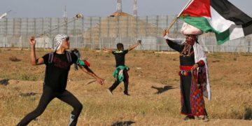 Un terrorista lanza piedras a las tropas israelíes mientras una mujer vestida con un traje tradicional palestino ondea una bandera nacional, durante los ataques cerca de la frontera con Israel, al este de la ciudad de Gaza, el 10 de mayo de 2019. (Dijo KHATIB / AFP)