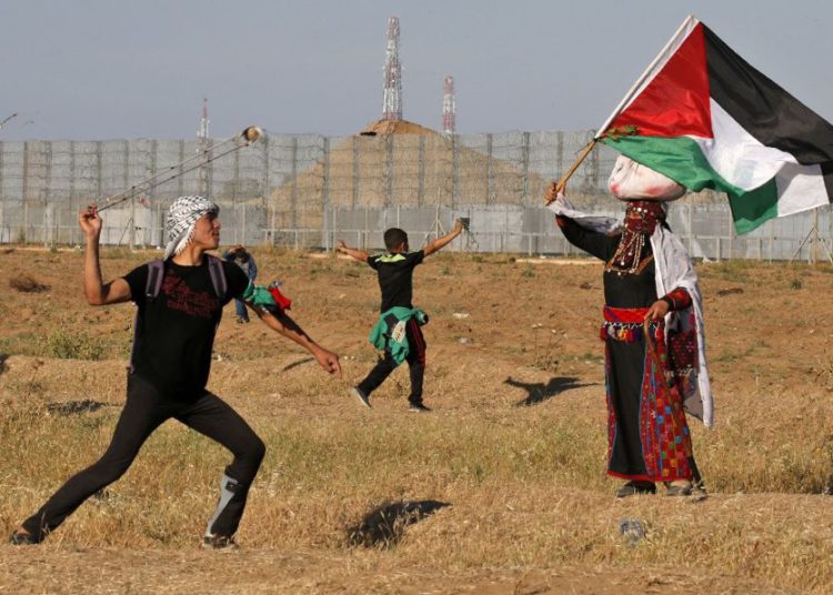 Un terrorista lanza piedras a las tropas israelíes mientras una mujer vestida con un traje tradicional palestino ondea una bandera nacional, durante los ataques cerca de la frontera con Israel, al este de la ciudad de Gaza, el 10 de mayo de 2019. (Dijo KHATIB / AFP)