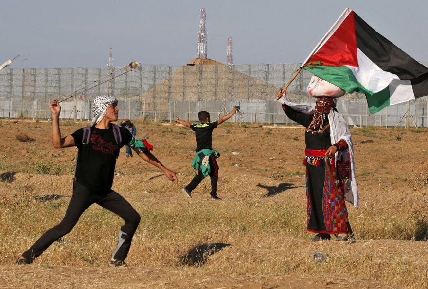 Un terrorista lanza piedras a las tropas israelíes mientras una mujer vestida con un traje tradicional palestino ondea una bandera nacional, durante los ataques cerca de la frontera con Israel, al este de la ciudad de Gaza, el 10 de mayo de 2019. (Dijo KHATIB / AFP)