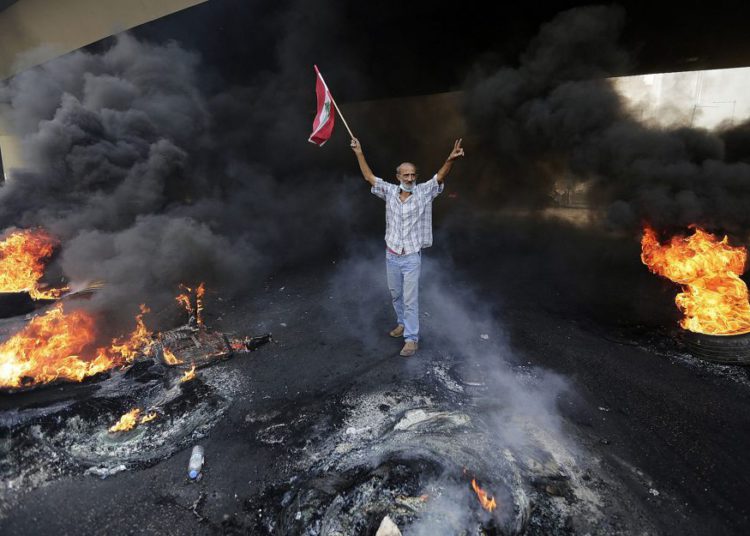 Un manifestante antigubernamental hace la señal de la victoria, mientras sostiene una bandera nacional libanesa y camina quemando neumáticos bloqueando una carretera durante una protesta contra los planes del gobierno de imponer nuevos impuestos en Beirut, Líbano, 18 de octubre de 2019. (Foto AP / Hassan Ammar )