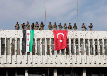 Soldados turcos, derecha y combatientes de la oposición respaldados por Turquía, izquierda, se paran en un edificio junto a sus banderas en la ciudad de Ras al Ayn, en el noreste de Siria, el 23 de octubre de 2019. (Ugur Can / DHA a través de AP)