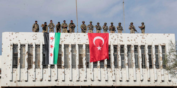 Soldados turcos, derecha y combatientes de la oposición respaldados por Turquía, izquierda, se paran en un edificio junto a sus banderas en la ciudad de Ras al Ayn, en el noreste de Siria, el 23 de octubre de 2019. (Ugur Can / DHA a través de AP)