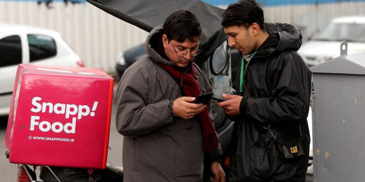 Un mensajero de comida para entregar bicicletas está junto a otro hombre mientras ambos usan teléfonos inteligentes a lo largo de una calle en la capital iraní, Teherán, el 23 de noviembre de 2019. (Atta Kenare / AFP)
