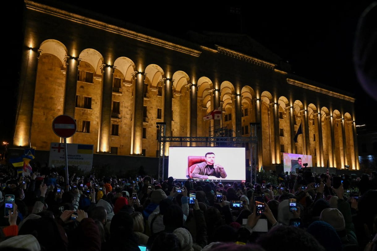 Manifestantes sostienen sus teléfonos y ven un discurso del presidente ucraniano Volodymyr Zelensky en la pantalla grande durante una manifestación en apoyo a Ucrania en Tiflis el 4 de marzo de 2022. (Vano SHLAMOV / AFP)