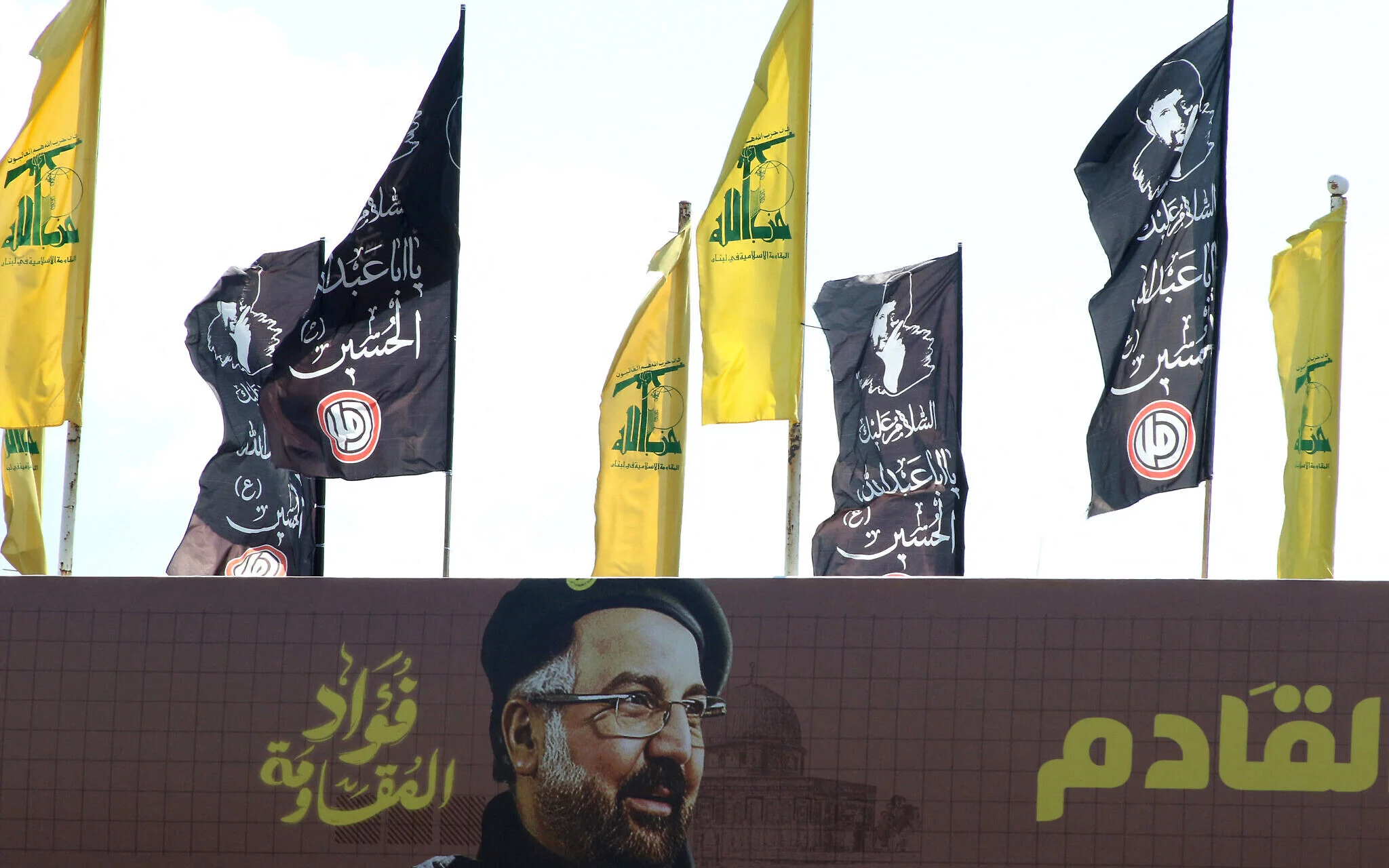 Banderas amarillas de Hezbolá y banderas negras de luto se izan junto a una pancarta que muestra al comandante militar del grupo terrorista, Fuad Shukr, quien fue asesinado en un ataque israelí, en una pasarela que cruza la carretera de Sidón a Tiro, en el sur del Líbano, el 2 de agosto de 2024. (Mahmoud Zayyat/AFP)