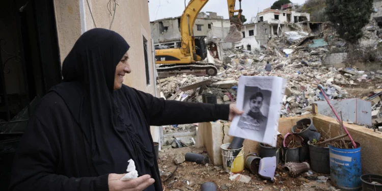 Una mujer sostiene un retrato de su nieto, un combatiente de Hezbolá que murió en los combates con las tropas israelíes, mientras espera que los rescatistas recuperen su cuerpo de debajo de los escombros de una casa destruida, al fondo, en la aldea de Ainata, en el sur del Líbano, tras un alto el fuego entre Israel y Hezbolá el 27 de noviembre de 2024. (Foto AP/Hussein Malla)