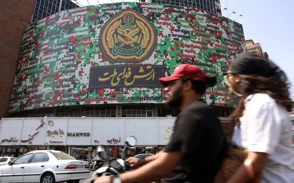 Los iraníes pasan junto a una valla publicitaria con los colores de la bandera nacional de Irán y llevan una frase en farsi: “El ejército se sacrifica por la nación” en la plaza Valiasr de Teherán, el 29 de agosto de 2025 (AFP) 