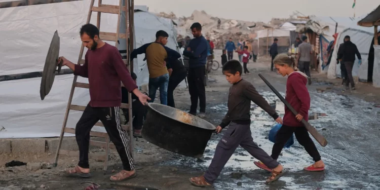 Palestinos desplazados caminan por senderos fangosos mientras un hombre y sus hijos cargan utensilios de cocina en un campamento en Jabalia, en el norte de la Franja de Gaza, el 7 de diciembre de 2025. (Foto AP/Jehad Alshrafi)