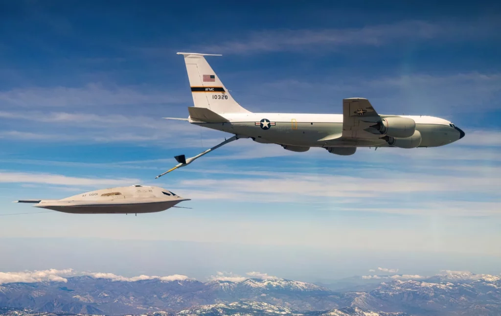 Dentro de su campaña de ensayos en la Base Aérea de Edwards, la Fuerza Aérea de EE. UU. confirmó el 14 de abril de 2026 que el B-21 Raider completó con éxito operaciones de reabastecimiento en vuelo. El avance refuerza la capacidad de ataque de largo alcance del nuevo bombardero estratégico y marca un paso relevante antes de su futura entrada en servicio dentro de la estructura de disuasión estadounidense. Las pruebas, ejecutadas junto con Northrop Grumman, forman parte del programa de evaluación con el que Washington busca validar la resistencia, el alcance global y la envolvente de rendimiento de la aeronave. Según la información oficial difundida con imágenes de los ensayos, la integración del reabastecimiento en vuelo busca reducir riesgos técnicos y acelerar el desarrollo sin renunciar a la madurez del sistema antes de su incorporación operativa. En el centro de esta fase aparece el KC-135 Stratotanker, el avión cisterna empleado para transferir combustible al Raider durante vuelos de larga duración. Ese aparato puede mover más de 90.000 kilogramos de combustible, según la configuración de la misión, y extiende la permanencia en el aire para sostener perfiles de ataque de largo alcance. En el B-21, la conexión se efectúa mediante un receptáculo dorsal ubicado detrás de la cabina. Las imágenes divulgadas muestran una compuerta de dos piezas tipo concha en esa zona, una solución distinta al sistema giratorio del B-2 Spirit. Esa disposición apunta a cambios dirigidos a reducir la firma de radar sin sumar complejidad mecánica. El avance llega además en un programa de pruebas y desarrollo concurrentes, mientras la Fuerza Aérea subraya el uso de ingeniería digital y procesos modernos de producción para acortar plazos de entrega. Durante esta fase de pruebas, las fotografías difundidas también revelan detalles de la configuración del bombardero. En ellas aparecen puertos de datos de aire delante de la cabina, característicos de la instrumentación de vuelos iniciales, además de tomas de aire con una geometría que parece optimizada para equilibrar sigilo y eficiencia de los motores. Esos rasgos refuerzan la orientación técnica del programa hacia un perfil de baja observabilidad. El B-21 retoma la arquitectura de ala volante introducida por el B-2, pero incorpora mejoras en el modelado de la sección transversal radar, materiales absorbentes de radar actualizados y estructuras compuestas. Su tamaño, ligeramente menor, apunta a reducir costos y mejorar la maniobrabilidad sin sacrificar la baja observabilidad frente a un amplio espectro de radares. La Fuerza Aérea sostiene además que consume bastante menos que el B-1B Lancer y el B-52H Stratofortress. Esa reducción de consumo, según funcionarios estadounidenses, tiene un efecto directo sobre el empleo operativo porque disminuye la dependencia de una flota limitada de aviones cisterna y libera más recursos de reabastecimiento para otros teatros. La aeronave estaría propulsada por dos motores optimizados para el sigilo, probablemente derivados de la familia Pratt & Whitney F-135, con un empuje superior a 11.000 kilogramos por unidad y firmas infrarroja y radar contenidas. Con esa configuración, el aparato tendría una velocidad de crucero cercana a 870 kilómetros por hora y una máxima próxima a 1.050 kilómetros por hora. Su alcance estimado ronda los 12.000 kilómetros y la carga útil se sitúa entre 12 y 13 toneladas. Con esas cifras, el B-21 ha sido concebido para ejecutar misiones intercontinentales con elevada autonomía, en coherencia con operaciones de penetración en entornos disputados y con su papel en la disuasión estratégica. En el plano del armamento, el bombardero mantendrá un doble cometido convencional y nuclear. Podrá transportar bombas gravitatorias nucleares B61 y B83, además del misil de crucero nuclear Long Range Stand Off. Para misiones convencionales, se prevé su compatibilidad con JDAM, JASSM y la GBU-57 Massive Ordnance Penetrator, destinada al ataque contra objetivos enterrados y fuertemente protegidos dentro de escenarios de alta complejidad defensiva. Las bodegas internas también han sido concebidas para admitir sistemas futuros, entre ellos armas hipersónicas, dentro de un enfoque centrado en la adaptabilidad. Esa lógica alcanza igualmente a la aviónica y al software. El B-21 integra fusión multisensor para combinar datos de radar, infrarrojos y guerra electrónica en un único cuadro operativo, mientras su arquitectura abierta permitiría incorporar nuevas capacidades con menos modificaciones estructurales a lo largo de su vida útil. La suite de guerra electrónica del Raider ha sido diseñada para interferir y degradar sistemas de defensa antiaérea enemigos, incluidas redes modernas de misiles tierra-aire, con el objetivo de aumentar la supervivencia del avión en escenarios de alta amenaza. En paralelo, el componente industrial del programa gana ritmo: la Fuerza Aérea mantiene como meta oficial una flota de al menos 100 aparatos y la capacidad de producción aumentó en torno a un 25 por ciento. El calendario vigente sitúa la capacidad operativa inicial en torno a 2027. Las primeras aeronaves destinadas al servicio deberían llegar a la Base Aérea de Ellsworth, en Dakota del Sur, donde ya se ejecutan trabajos de infraestructura y ajustes de pista e instalaciones para sistemas de baja observabilidad. Frente al número reducido de B-2 Spirit y a la falta de características furtivas del B-52H, el Raider ampliará las opciones de ataque de largo alcance de EE. UU.