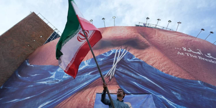 Un hombre ondea una bandera iraní en apoyo a una campaña progubernamental bajo una valla publicitaria con una imagen del estrecho de Ormuz y los labios cosidos del presidente estadounidense Donald Trump en una plaza del centro de Teherán, el 6 de mayo de 2026. (Foto AP/Vahid Salemi)