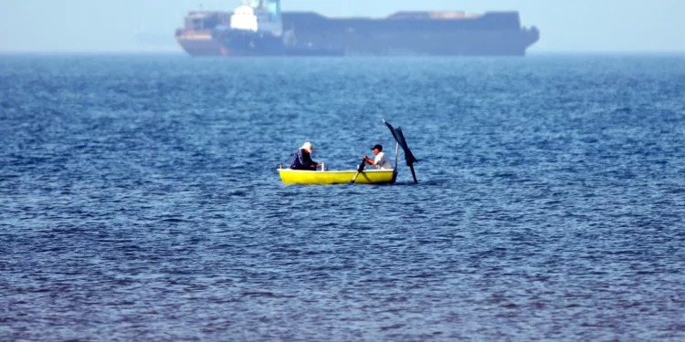 En esta imagen obtenida de la agencia de noticias iraní ISNA el 4 de mayo de 2026, se ve a dos hombres sentados en una barca pescando cerca de un buque anclado en el estrecho de Ormuz, frente a Bandar Abbas, en el sur de Irán. (Amirhossein KHORGOOEI / ISNA / AFP)