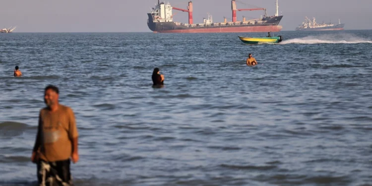 Unos residentes se bañan en las aguas del estrecho de Ormuz mientras una pequeña lancha a motor rebasa a buques de carga y otras embarcaciones comerciales en alta mar, cerca de Bandar Abbas (Irán), el 17 de junio de 2026. (Amirhosein Khorgooi/ISNA vía AP)