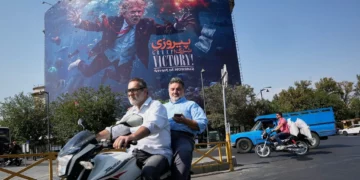 Motociclistas pasan junto a un cartel con Donald Trump bajo el agua y explosiones en Teherán, el 24 de agosto de 2026.