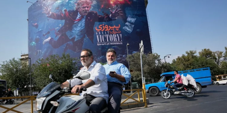 Motociclistas pasan junto a un cartel con Donald Trump bajo el agua y explosiones en Teherán, el 24 de agosto de 2026.