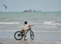 Un niño con una bicicleta observa buques mercantes fondeados en el estrecho de Ormuz mientras una gaviota vuela sobre la playa.