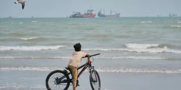 Un niño con una bicicleta observa buques mercantes fondeados en el estrecho de Ormuz mientras una gaviota vuela sobre la playa.